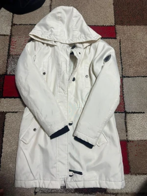 Vit parkas med huva och fickor - Snygg vit parkas med huva och dragkedja framtill. Jackan har två stora fickor med lock, svarta detaljer vid ärmsluten och en patch på ena ärmen. Lång modell som passar perfekt när det är kallt ute. Stängs med både dragkedja och knappar.Jacka  Pälsdetaljen på huvan saknas.