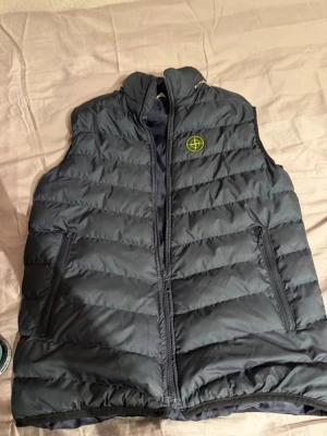Mörkgrå dunväst med logga - Snygg mörkgrå dunväst med quiltad design och dragkedja framtill. Västen har två praktiska fickor med dragkedja och en broderad logga på bröstet. Perfekt för lager-på-lager och chill stil.