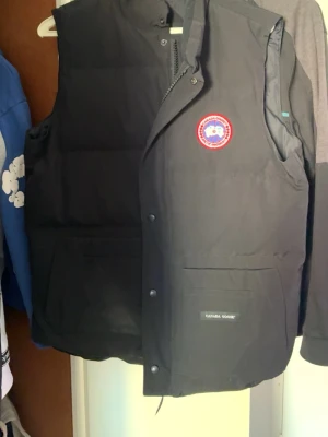 dunväst från Canada Goose äkta  - Snygg svart dunväst från Canada Goose med klassisk logga på bröstet och två stora fickor framtill. Västen har tryckknappar och dragkedja samt diskret Canada Goose-märke på ena fickan. Perfekt för lager-på-lager och håller dig varm under kyliga dagar.