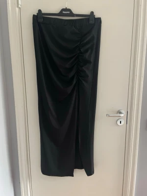 Svart långkjol med drapering - Snygg svart långkjol i satinliknande material med draperad detalj framtill och slits. Kjolen har en rak passform och ger en elegant look. Perfekt att styla med både sneakers och klackar.