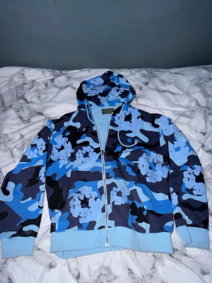 Blå camo hoodie från Denim Tears - Säljer en riktigt snygg hoodie från Denim Tears i blått camo-mönster med vita blommor. Hoodien har dragkedja, huva med dragsko och ribbade muddar. Materialet är mjukt och skönt, perfekt för chill dagar eller streetwear-stil. Pris går att diskutera