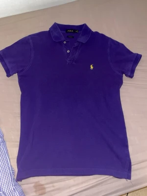 Lila pikétröja från Polo Ralph Lauren - Säljer en klassisk lila pikétröja från Polo Ralph Lauren med gul broderad logga på bröstet. Tröjan har krage, knappar vid halsen och korta ärmar. Perfekt för en avslappnad men ändå stilren look.