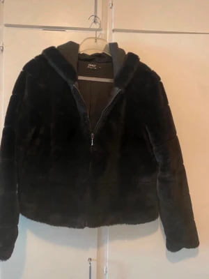 Svart teddyjacka med huva - Mysig svart teddyjacka från ONLY med dragkedja framtill och stor huva. Jackan har en fluffig, mjuk yta och två sidofickor. Perfekt för kyliga dagar när du vill vara både varm och snygg.