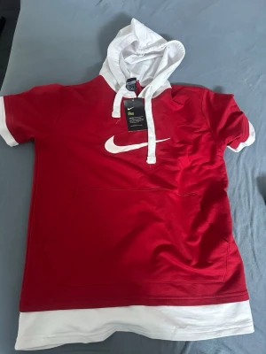 Röd Nike hoodie med vit huva - Snygg röd hoodie från Nike med vit huva och vita detaljer på ärmar och nederkant. Stor vit Nike-logga på bröstet och känguruficka framtill. Kortärmad modell i funktionsmaterial med Dri-FIT-teknologi som håller dig torr och bekväm.