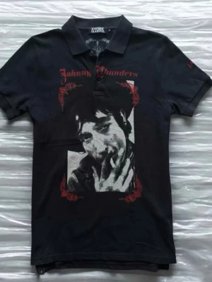 hysteric glamour polo - Säljer en svart t-shirt med krage och tryck av Johnny Thunders på framsidan. T-shirten har korta ärmar, knappar vid halsen och röda detaljer runt motivet. Perfekt för dig som gillar rockig stil och vill sticka ut.