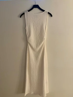 Vit ärmlös långklänning - Stilren vit långklänning med ärmlös design och rund halsringning. Klänningen har en lätt strukturerad yta och rak passform som ger en enkel men elegant look. Perfekt för sommarens alla tillfällen.