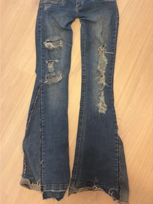 Virala jeans - Säljer ett par blå bootcut jeans med rejält slitna detaljer och hål på både fram- och baksida. Jeansen har fransiga benslut och klassisk femficksmodell. Materialet är denim med synliga sömmar i kontrastfärg. Perfekta för dig som gillar en edgy och avslappnad stil.