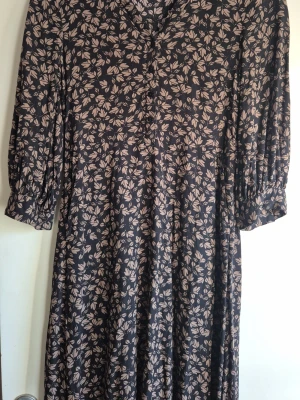 Svart långklänning med blommigt mönster - Mönstrad svart långklänning med beige blommor och trekvartsärmar. Klänningen har en avslappnad passform och knappar framtill. Perfekt för dig som gillar en stilren men ändå söt look.