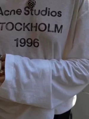 Vit långärmad Acne Studios tröja - Vit långärmad topp från Acne Studios med tryck i svart text 'STOCKHOLM 1996' på bröstet. Tröjan har avslappnad passform, rund halsringning och är gjord i mjukt bomullsmaterial. Perfekt för en clean och trendig look.