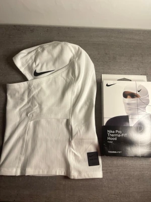 Nike Pro Therma-FIT vit balaclava - Nike Pro Therma-FIT Hood i vitt, tillverkad i ett stretchigt och värmande material. Balaclavan har svart Nike-logga på sidan och en diskret etikett nedtill. Perfekt för träning utomhus när det är kallt.