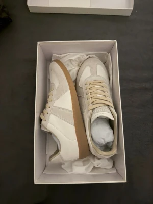 Masion Margiela gats - Maison Margiela sneakers vit, mocka med beige detaljer och snörning. Klassisk rund tå och platt gummisula i brunt. Är du intresserad av fler bilder eller vill veta mer, är det bara att höra av dig!