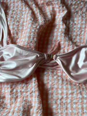 Ljusrosa bikiniöverdel med knyt - Supersöt ljusrosa bikiniöverdel med tunna axelband och knyt både fram och bak. Bikinin har en enkel och stilren design med lätt glansigt material och en liten gulddetalj framtill. Perfekt för lata dagar på stranden eller poolen.
