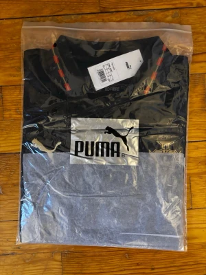 Svart och grå Puma t-shirt - Snygg svart och grå t-shirt från Puma med stor logga på bröstet. T-shirten har rund halsringning med röda detaljer och är tillverkad i ett mjukt material. Perfekt för dig som gillar sportig stil och vill ha något bekvämt och trendigt.