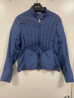 Blå vindjacka från Colmar - Snygg blå vindjacka från Colmar med quiltade detaljer och hög krage. Jackan har dragkedja framtill, två fickor och Colmar-logga på ärmen. Perfekt för blåsiga dagar och enkel att matcha med olika outfits.