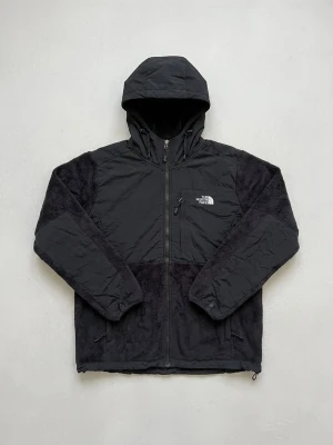 Svart teddyjacka med huva The North Face - Snygg svart teddyjacka från The North Face med huva och dragkedja framtill. Jackan har mjuk teddyfleece på ärmar och nederdel samt slät yta upptill. Logga på bröstet och ryggen. Perfekt för kalla dagar och riktigt skön att bära.