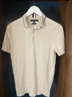 Beige merinoullsskjorta Tommy Hilfiger - Snygg beige skjorta från Tommy Hilfiger i mjuk merinoull. Skjortan har korta ärmar, klassisk krage med kontrastkant och två knappar framtill. Perfekt för dig som gillar stilrena och bekväma plagg.