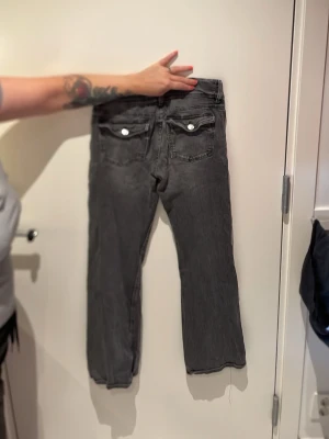 Grå raka jeans med fickdetaljer - Säljer ett par grå jeans med utsvängda ben. Jeansen har knappar på bakfickorna och en snygg tvättad look. Passar perfekt till en avslappnad stil och är tillverkade i denim.