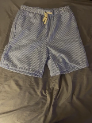 Blåvitrandiga shorts med snörning - Säljer ett par blåvitrandiga shorts med elastisk midja och vit snörning framtill. Shortsen har sidofickor och är tillverkade i ett lätt och luftigt bomullstyg, perfekta för varma dagar.