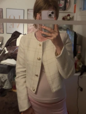 Vit tweedjacka med guldknappar - Stilren vit tweedjacka med diskret rutmönster och raka axlar. Jackan har rund halsringning och stängs med tre dekorativa guldknappar framtill. Kort, boxig modell som ger en chic look och passar perfekt till både jeans och kjol.