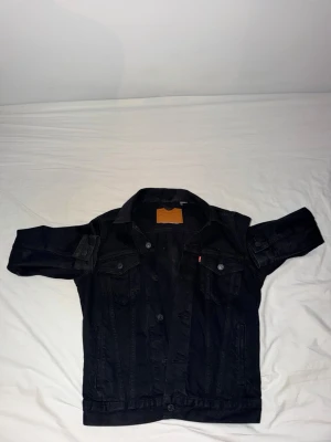 Svart jeansjacka från Levis med klassisk look - Snygg svart jeansjacka från Levis  med raka linjer och klassisk krage. Jackan har två bröstfickor med knappar och knappstängning framtill. Passar perfekt till en avslappnad streetstil och är enkel att matcha med olika outfits.