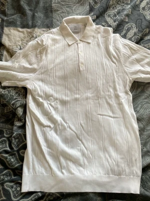 Vit polotopp från Bläck i M - Vit polotopp från Bläck i storlek M. Toppen har klassisk krage, knappar framtill och diskreta vertikala ränder i strukturen. Perfekt för en clean och stilren look. Materialet känns mjukt och bekvämt mot huden.