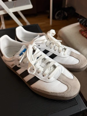 Adidas Samba vita och svarta sneakers - Klassiska Adidas Samba sneakers i vitt läder med svarta ränder och grå mockadetaljer på tån. Skorna har vit sula och vita skosnören samt blå Adidas-loggan på plösen. Perfekta för en stilren och sportig look. Använda ca 3ggr utomhus så de är i fint skicka! Nypris 1500kr