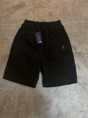 Svarta shorts från Polo Ralph Lauren - Svarta shorts från Polo Ralph Lauren med elastisk midja och snörning. Klassisk röd logga broderad på vänster ben. Sköna och stilrena, perfekta för sommaren. Materialet känns mjukt och bekvämt, troligen bomull. Den är i storlek L men funkar också om man brukar h storlek M den säljs billigt eftersom fick den i present och den var liten så den är bara testad 