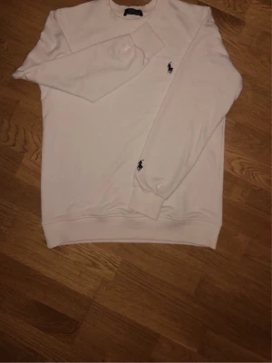 Vit sweatshirt från Polo Ralph Lauren - Klassisk vit sweatshirt från Polo Ralph Lauren med rund halsringning och broderad marinblå logga på bröstet och ärmen. Tröjan har ribbade muddar vid ärmslut och nederkant. Tillverkad i mjukt bomullsmaterial för en skön och avslappnad look.