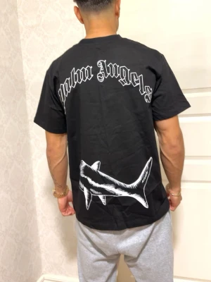 Svart Palm Angels t-shirt med hajmotiv - Svart t-shirt från Palm Angels med stor vit hajgrafik både fram och bak. T-shirten har korta ärmar och klassisk passform. Stort Palm Angels-tryck i vitt på ryggen. Perfekt för dig som gillar streetwear och coola prints.