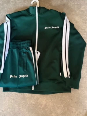 Grön tracksuit från Palm Angels - Mörkgrön tracksuit från Palm Angels med vita ränder längs ärmar och ben. Setet består av ziptröja och byxor, båda med logga broderad i vitt. Tröjan har hög krage och dragkedja, byxorna har resår i midjan och sidofickor. Perfekt för chill dagar.