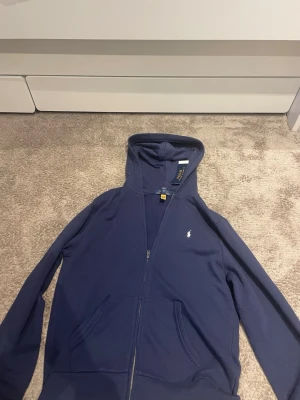 Blå hoodie från Polo Ralph Lauren - Säljer en blå hoodie från Polo Ralph Lauren med dragkedja och huva. Klassisk design med liten vit logga på bröstet och två fickor framtill. Tillverkad i mjukt bomullsmaterial, perfekt för en avslappnad och stilren look.