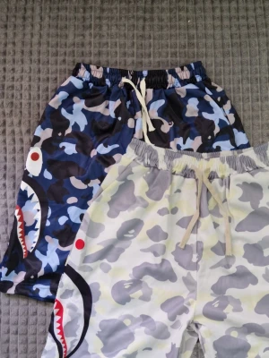 Två Bape shorts med hajmotiv och snörning - Två par Bape camo shorts med hajmotiv på sidan. Ena paret är blå, svart och vit camo, det andra är ljusgrå, vit och beige camo. Båda har snörning i midjan och elastisk midja för skön passform. Perfekta för sommaren och streetwear.