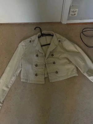 Beige kort militärinspirerad jacka - Stilren beige kort jacka med militärinspirerade detaljer och dekorativa knappar framtill. Jackan har axelklaffar och långa ärmar, perfekt för att lyfta en outfit med en edgy touch. Passar dig som gillar statement-plagg och vill sticka ut.