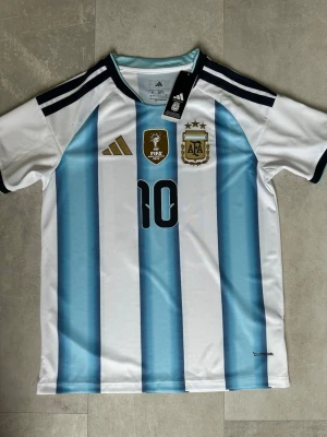 Argentina matchtröja nr 10 Adidas - Säljer en helt ny Argentina matchtröja från Adidas med klassiska blå och vita ränder, guldiga detaljer och Messi på ryggen. Om du vill ha en stilig fotbollströja så är denna den perfekta. 