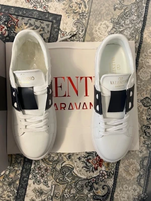 Vita Valentino Garavani sneakers - Snygga vita sneakers från Valentino Garavani med svart band och silverfärgade nitar på sidan. Skorna har en klassisk låg profil, vita snören och en tjock vit sula. Tillverkade i slätt läder för en clean och exklusiv look.