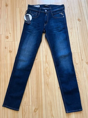 Mörkblå slim fit jeans Replay - Snygga mörkblå slim fit jeans från Replay, modell Anbass. Jeansen har klassisk femficksdesign, kontrastsömmar och knappgylf. Perfekta för dig som gillar en smalare passform och stilren look.