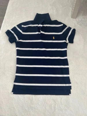 Marinblå randig pikétröja Ralph lauren  - Snygg marinblå pikétröja med vita horisontella ränder och klassisk krage. Tröjan har korta ärmar och en liten gul broderad logga på bröstet. Perfekt till jeans eller shorts för en avslappnad look.