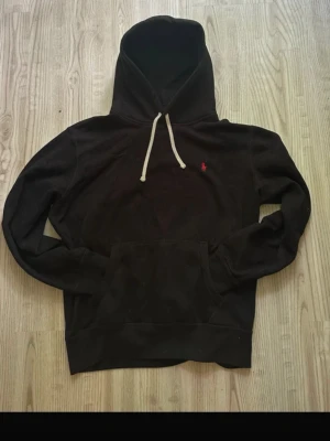 Svart ralph lauren hoodie med vita snören - Säljer en svart hoodie med vit dragsko och känguruficka. På bröstet finns en liten röd broderad logga. Hoodien har en klassisk passform och är tillverkad i mjukt material, perfekt för en avslappnad stil. Köptes på boostlet för 1200kr har helt änkelt blivit för stor.              KOM MED PRISFÖRSLAG