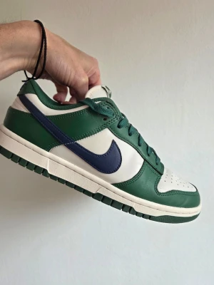 Nike Dunk Low grön/vit/blå - Snygga Nike Dunk Low sneakers i grönt och vitt med mörkblå swoosh. Skorna har klassisk låg siluett, perforerad tåbox och ovandel i läder. Yttersulan är grön och mellansulan vit. Perfekta för dig som gillar streetwear och sportig stil.