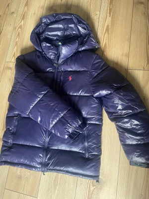 Mörklila pufferjacka med huva - Snygg mörklila pufferjacka från Ralph Lauren med glansig finish och klassisk röd logga på bröstet. Jackan har huva, dragkedja framtill och två sidofickor med dragkedja. Perfekt för kalla vinterdagar och riktigt trendig look, passar nog runt 180-190hållet, användt 2 gånger totalt, inga alls skador eller defekter, funkar precis som den ska , mer bilder kan skickas privat 