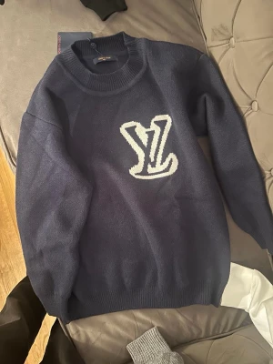 Mörkblå stickad tröja med vit logga - Mörkblå stickad tröja. Tröjan har rund halsringning, ribbstickade muddar vid ärmslut och nederkant samt en avslappnad passform. Perfekt för dig som gillar streetwear och vill ha något som sticker ut.
