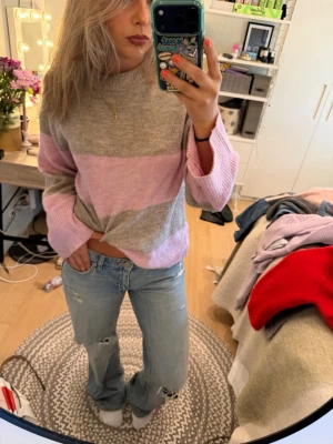 Stickad tröja med rosa och grå ränder - Mysig stickad tröja med breda horisontella ränder i ljusrosa och ljusgrått. Tröjan har en avslappnad passform, rund halsringning och långa ärmar. Perfekt för dig som gillar en chill och trendig stil.