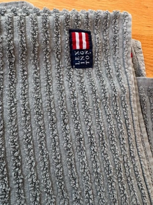 Grå Lexington handduk med struktur - Säljer en grå handduk från Lexington med tydlig ribbad struktur och en broderad etikett med Lexingtons logga i blått, vitt och rött. Handduken är mjuk och har en modern look som passar i de flesta badrum.