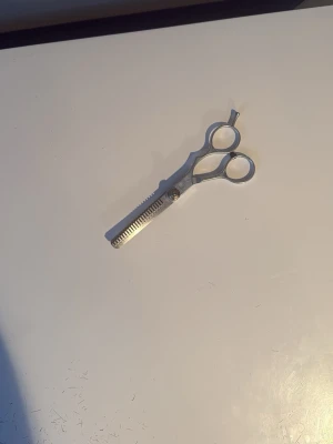Silvriga effileringssaxar för hår - Säljer ett par silvriga effileringssaxar i metall, perfekta för att tunna ut och forma håret. Saxarna har ett klassiskt grepp med två fingerhål och en tandad sida för att skapa mjuka övergångar i frisyren.