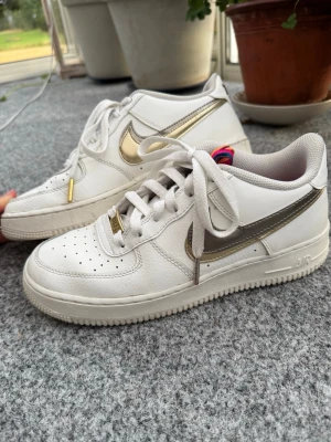 Vita Nike air force 1 sneakers med guld & silver detaljer - Snygga vita Nike air force 1 sneakers med metallic guld- & silverfärgad båge på sidorna!🤩Skorna har klassisk låg modell, vita snören med guld & silver detaljer! Samt perforerad tå för extra ventilation och guld/silver metallbricka vid snörena🙌 Perfekta för dig som gillar stilrena och trendiga sneakers! Dam storlek 40 (25cm). Använda flera gånger men inga stora fel, har tecken på användning då köpta secondhand🩷Så unika och annorlunda design (ungdomsskor), svåra att hitta/få tag på❤️