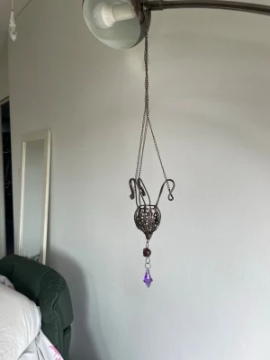 Dekorativ hängande smyckeshållare - Unik smyckeshållare i mörk metall med konstnärlig, burformad design. Hängs upp med kedja och har dekorativa detaljer samt en lila kristall-liknande pärla i botten. 