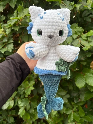 Virkad mermaid kitty💙 - Unik handgjort gosedjur i form av en katt-sjöjungfru. Virkad i mjukt plyschgarn och har vita och blå detaljer, gröna ögon och en blågrön fiskstjärt med dekorativa blad. Perfekt som present eller dekoration. Rekommenderar dock inte till småbarn då ögonen utgör en kvävningsrisk!