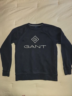 Mörkblå sweatshirt från GANT - Snygg mörkblå sweatshirt från GANT med vit logga och tryck på bröstet. Tröjan har rund halsringning, långa ärmar och ribbade muddar vid ärmslut och nederkant. Perfekt för en avslappnad och stilren look.