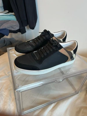 Svarta burberry skor  - Snygga svart burberry med beige och svart rutigt mönster på sidan. Skorna har vita sulor, svarta skosnören och en låg profil. Materialet ser ut att vara en mix av läder och textil, vilket ger en cool kontrast mellan olika ytor.
