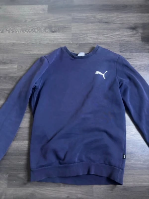 Mörkblå sweatshirt från Puma - Mörkblå sweatshirt med rund halsringning och lång ärm från Puma. Tröjan har en vit Puma-logga på bröstet och ribbade muddar vid ärmslut och nederkant. Perfekt för en avslappnad och sportig stil.